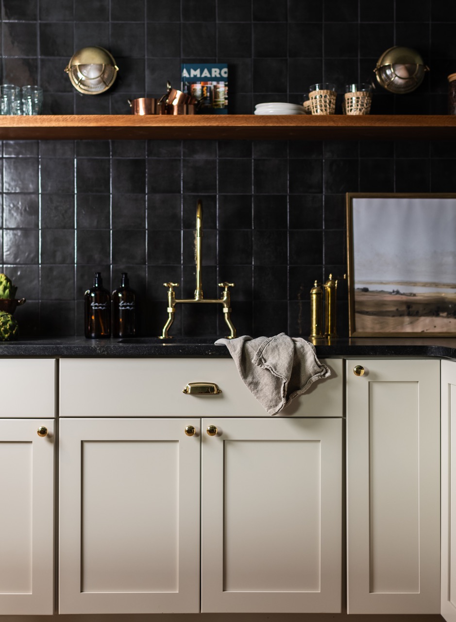 Modern kitchen sink area with brass faucet, black tiled backsplash, white cabinets, wooden shelf with glassware, and a beige towel draped over the sink.