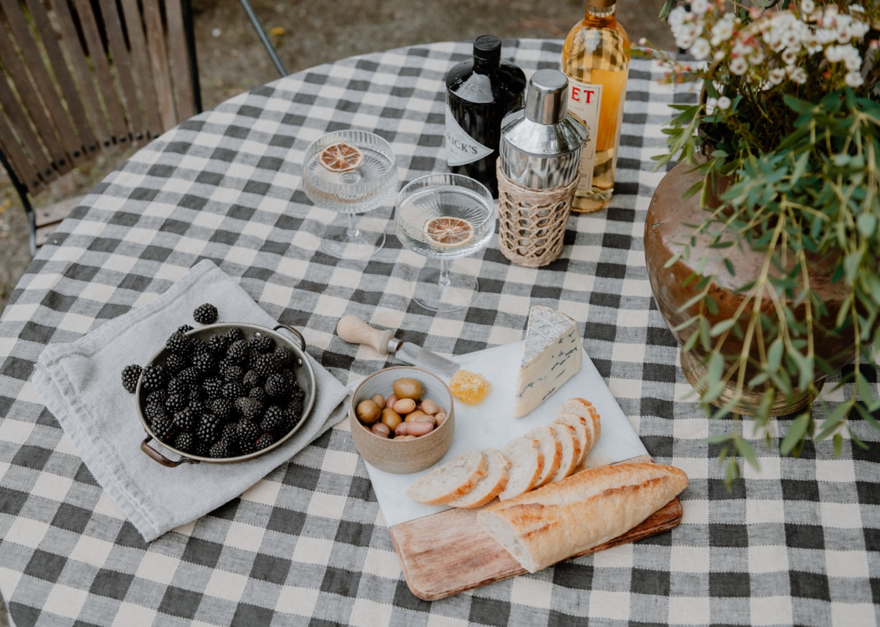 Checkered table with sliced baguette, blue cheese, olives, honeycomb, blackberries, cocktail bottles, two glasses with dried citrus garnish, and a large vase with flowers.