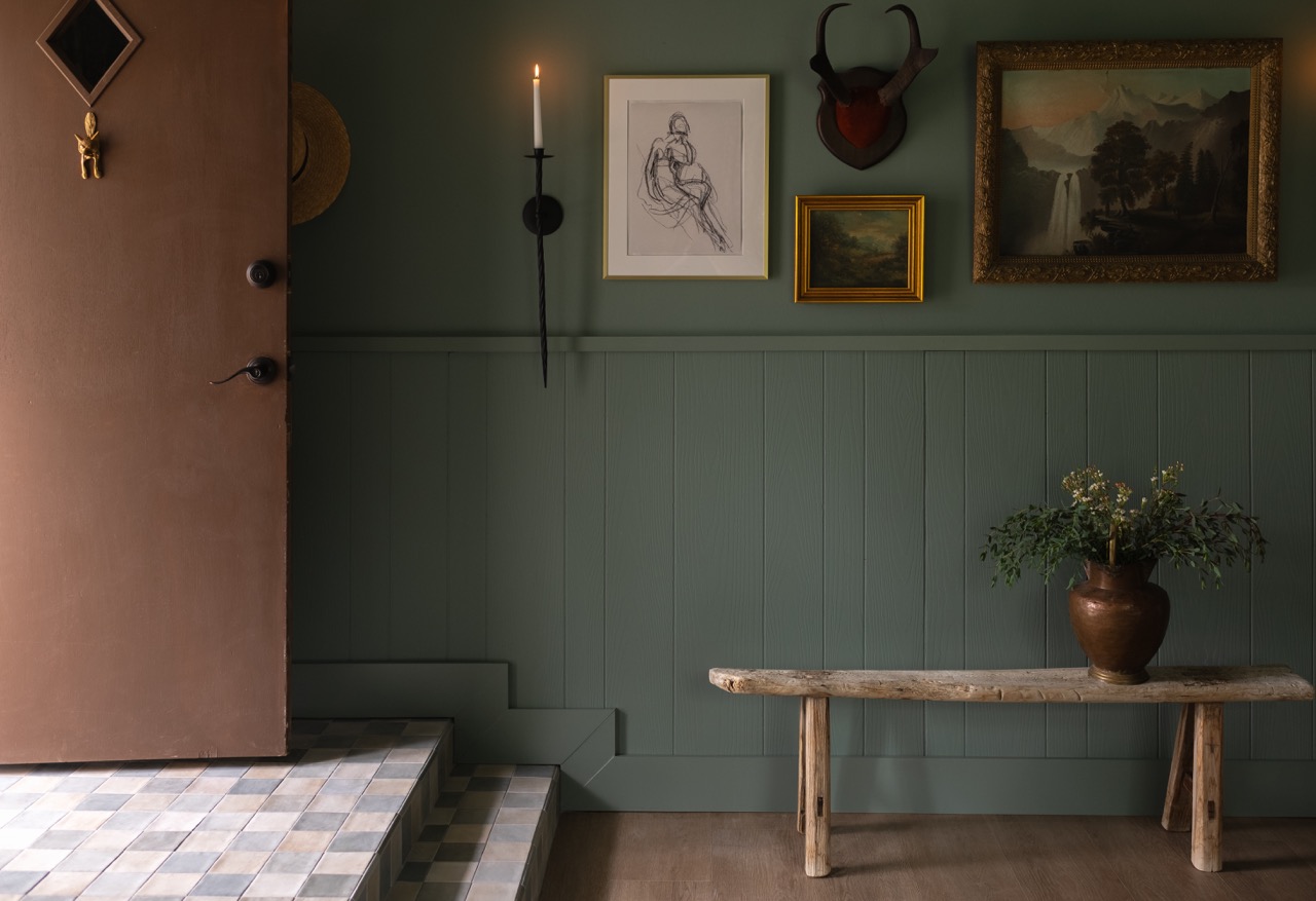 Interior entryway with a brown door, tiled steps, green paneled wall, wooden bench with vase of flowers, wall-mounted candle, framed sketches, and mounted antelope horns.
