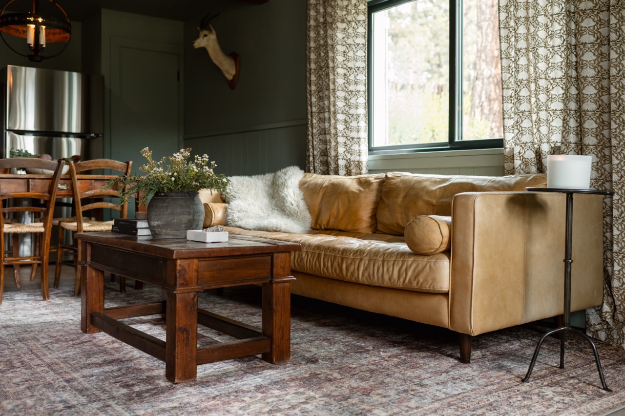 Cozy living room with a tan leather sofa, wooden coffee table with a potted plant, patterned curtains, and dining area in the background.