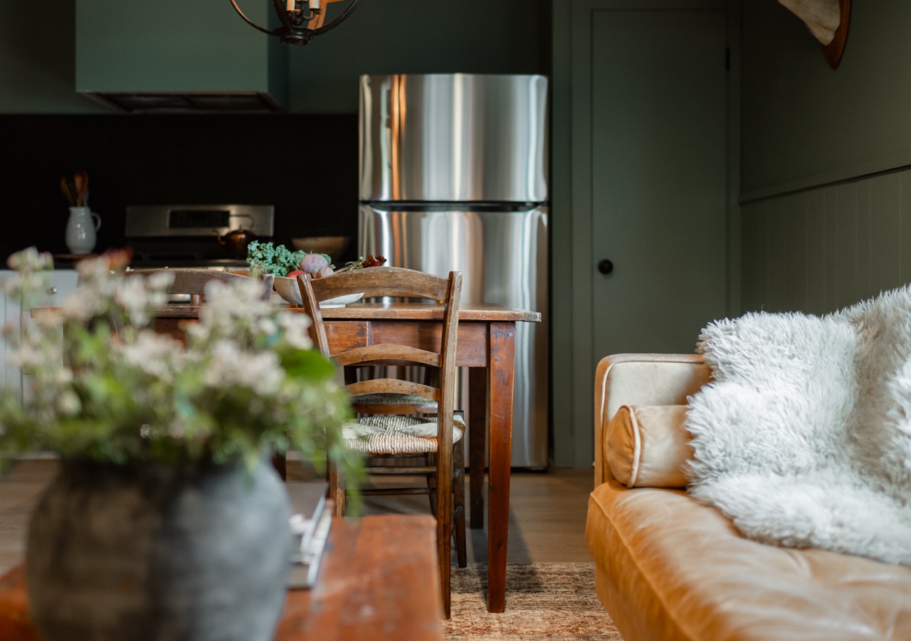 Cozy rustic kitchen and living area with wooden dining table, chairs, stainless steel refrigerator, and tan leather sofa with white fluffy pillow.