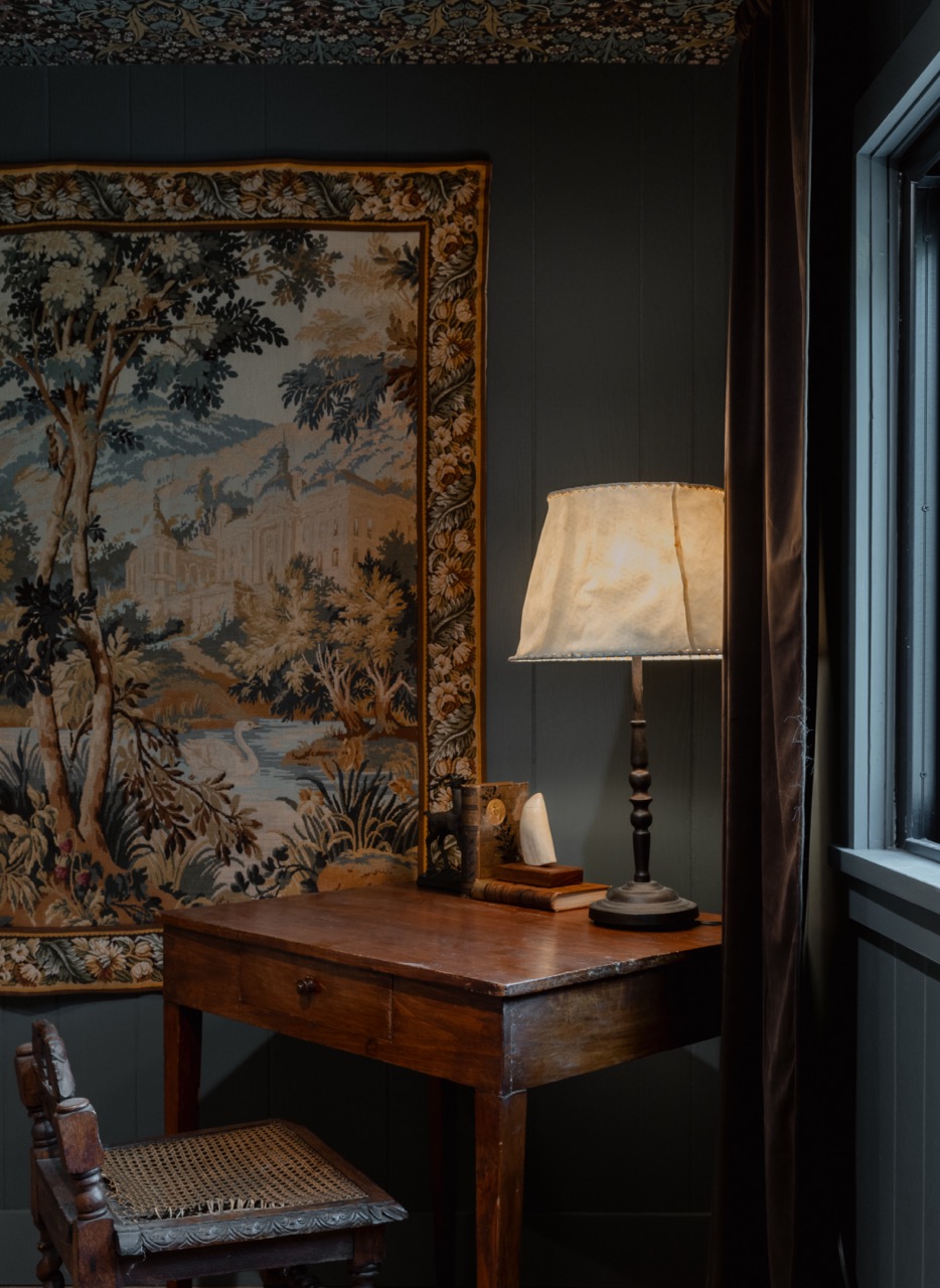 Wooden desk with a lit lamp, books, and a decorative item next to a vintage tapestry and a window with dark curtains.