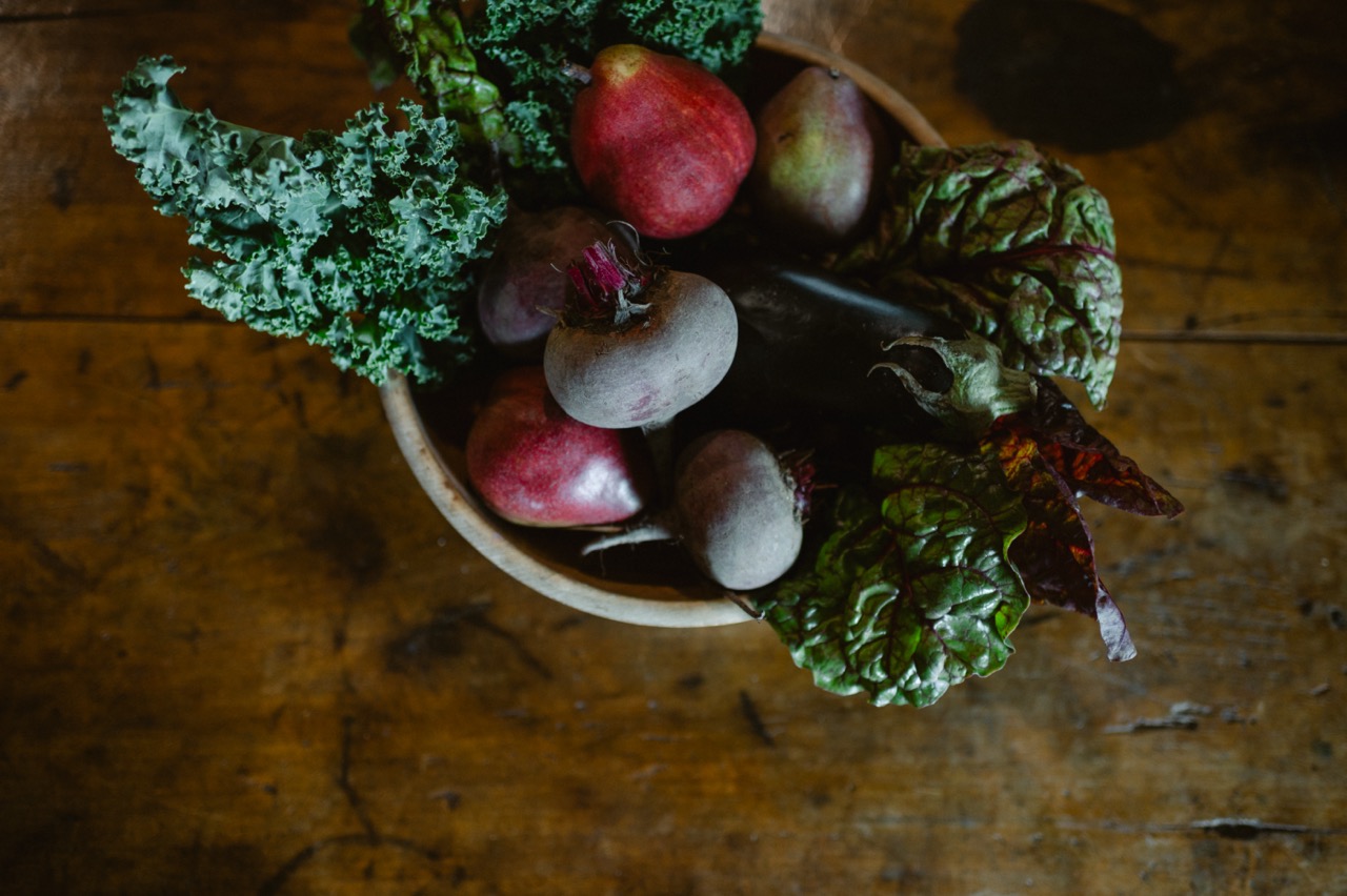Wooden bowl filled with fresh kale, beets, pears, eggplant, and leafy greens on a rustic wooden table.
