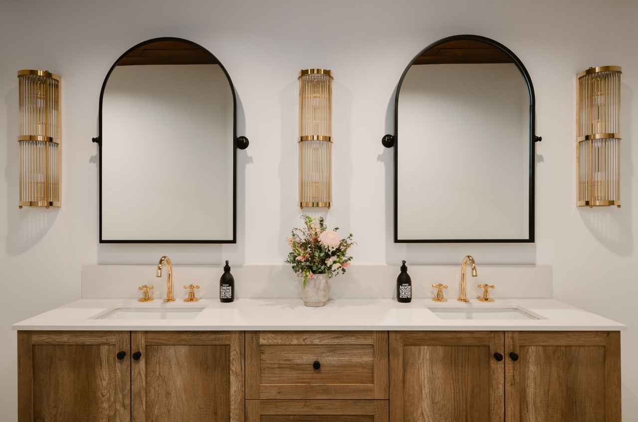 Modern double bathroom vanity with wooden cabinets, white countertop, gold faucets, black-framed arched mirrors, gold sconces, and a floral arrangement in the center.