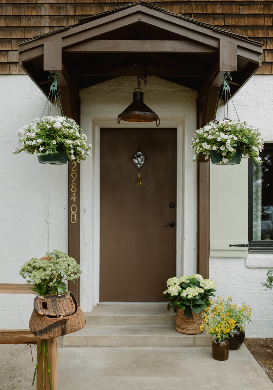 Front porch with brown door under wooden awning, hanging baskets with white flowers, and pots of green and yellow plants on concrete steps.