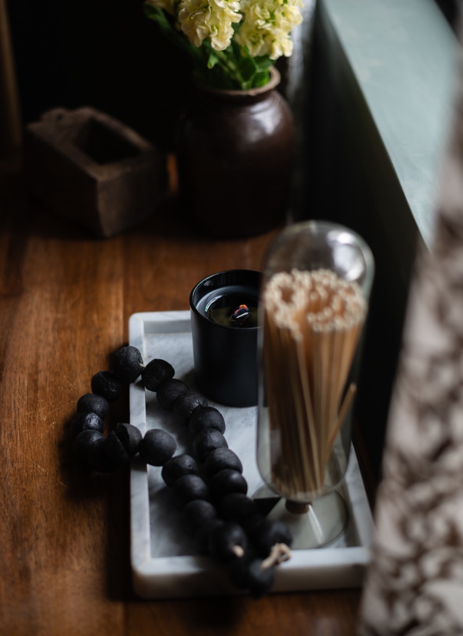 Black candle, wooden beads, and covered matches arranged on a marble tray on a wooden surface.