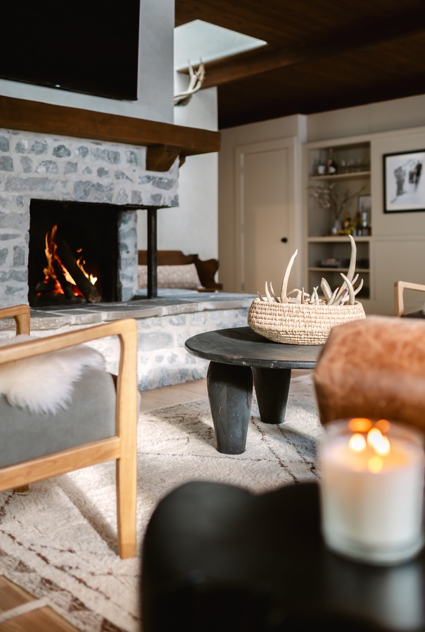 Cozy living room with a lit stone fireplace, wooden furniture, black coffee table with a woven basket of antlers, and a lit candle in the foreground.