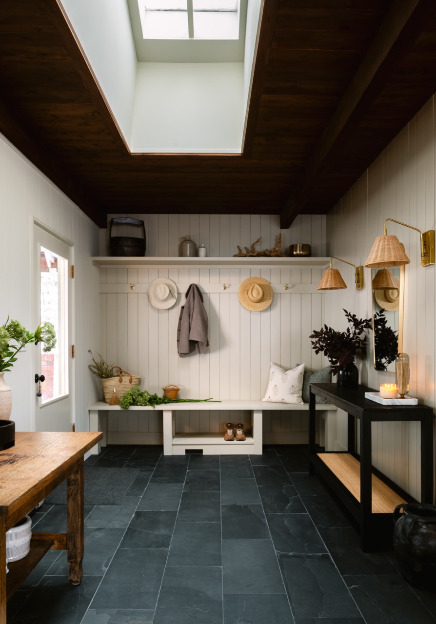 Bright entryway with white wood panel walls, dark slate floor tiles, skylight, bench with hats and coat hooks, and black console table with candles and plants.
