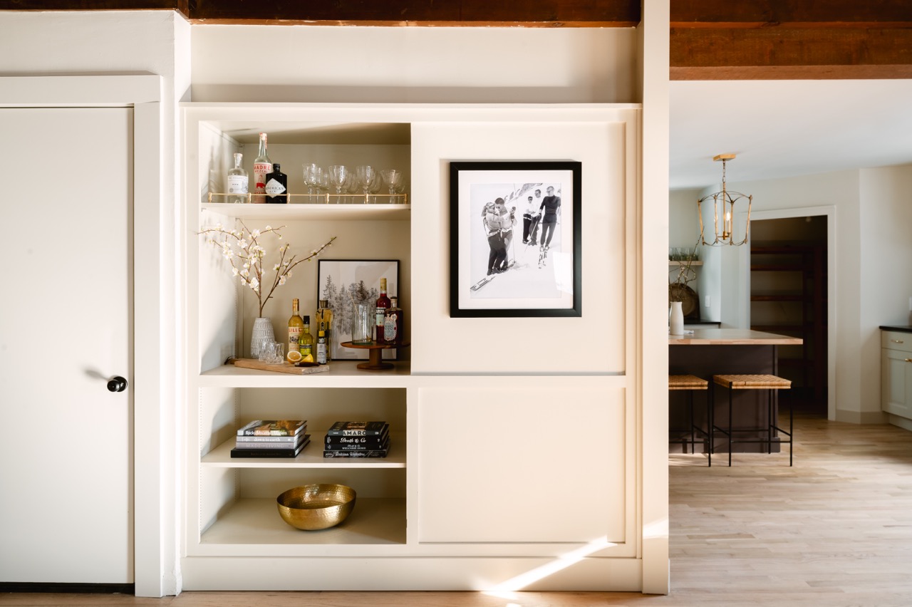 Built-in cream bookshelf with decorative bottles, glasses, framed photos, books, and a gold bowl beside a kitchen area with wooden stools and a hanging light fixture.