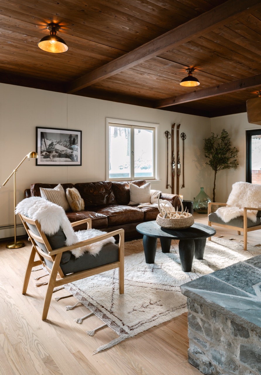 Cozy living room with wooden ceiling, leather sofa, two armchairs with white fur throws, a round black coffee table, and vintage skis on the wall.