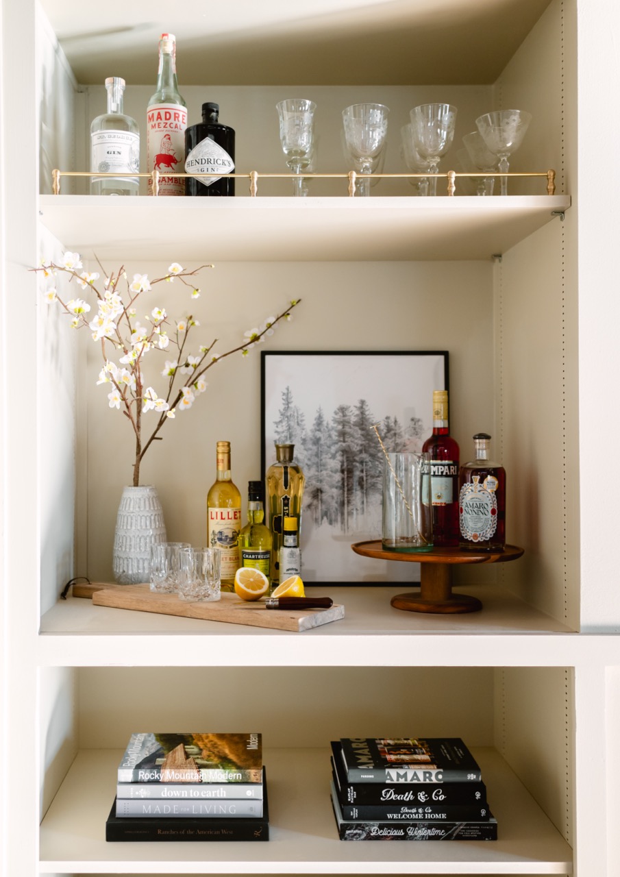 Shelf with bottles of liquor, glassware, a vase with white flowers, lemons on a cutting board, and a framed forest artwork, with stacks of books below.
