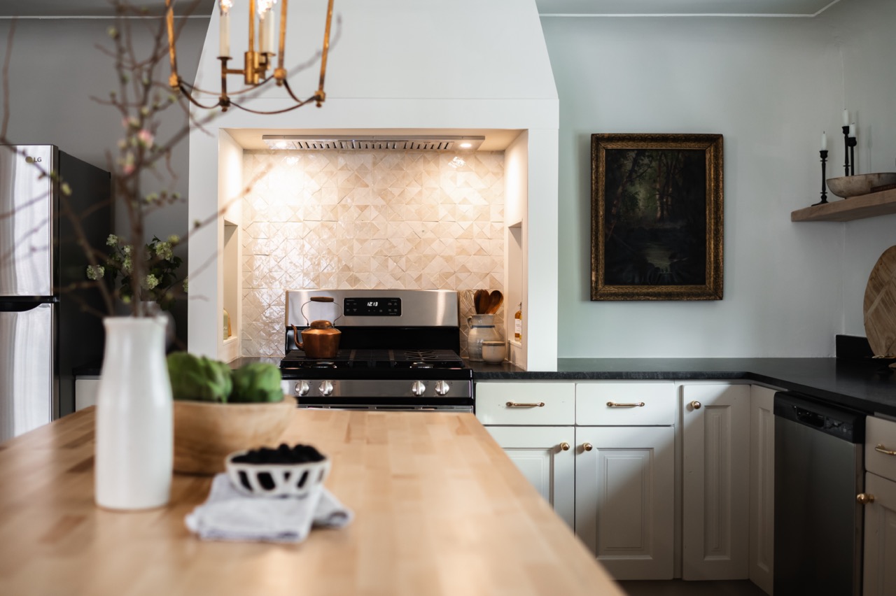 Modern kitchen with white cabinets, black countertops, stainless steel appliances, patterned tile backsplash, and a wooden table with a vase and fruits.