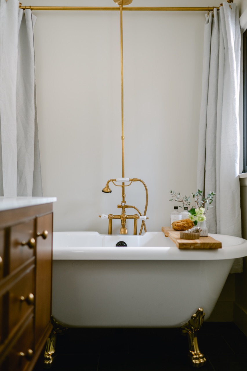 White freestanding clawfoot bathtub with gold fixtures and a wooden tray holding a loofah and small vase, set against a light wall with white curtains.