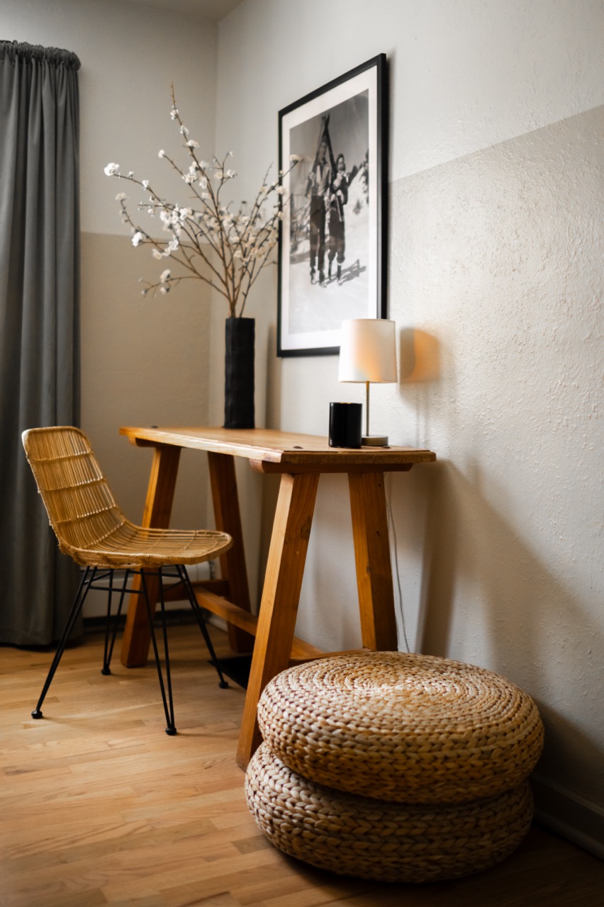 Cozy minimalist workspace with a wooden desk, wicker chair, table lamp, black vase with white flowers, and two stacked woven cushions on a wood floor.