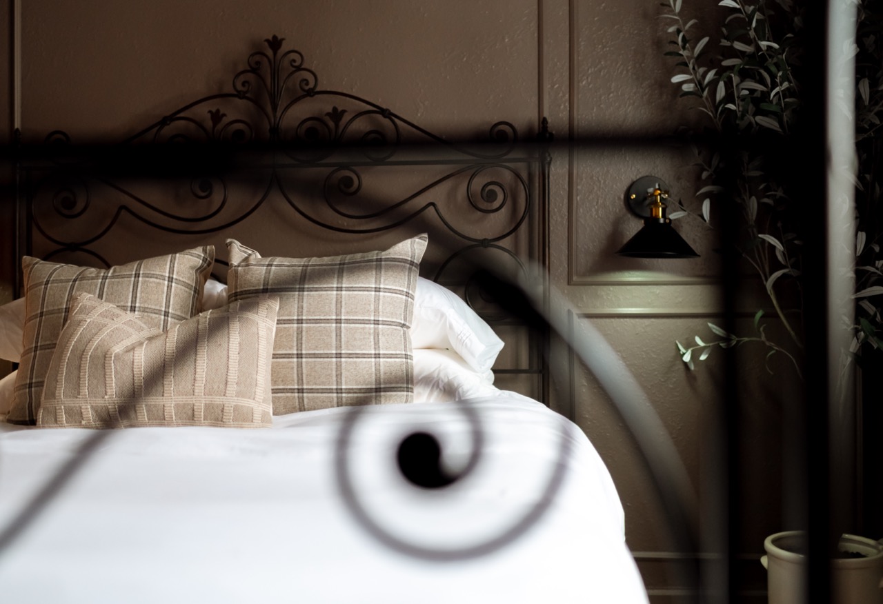 Bed with white linens, beige patterned pillows, and a decorative black wrought iron headboard, with a wall lamp and potted plant beside it.