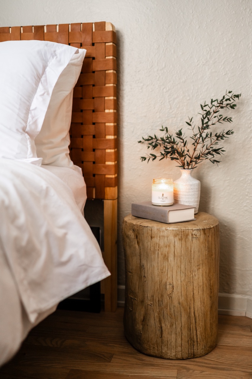Rustic wooden bedside table with a gray book, lit Old Pine scented candle, and white vase with green branches next to a bed with a woven leather headboard.