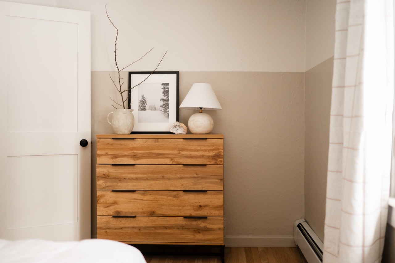 Minimalist wooden dresser with a ceramic vase holding bare branches, a framed black-and-white tree photo, a round lamp, and a decorative stone on top in a neutral-toned bedroom.