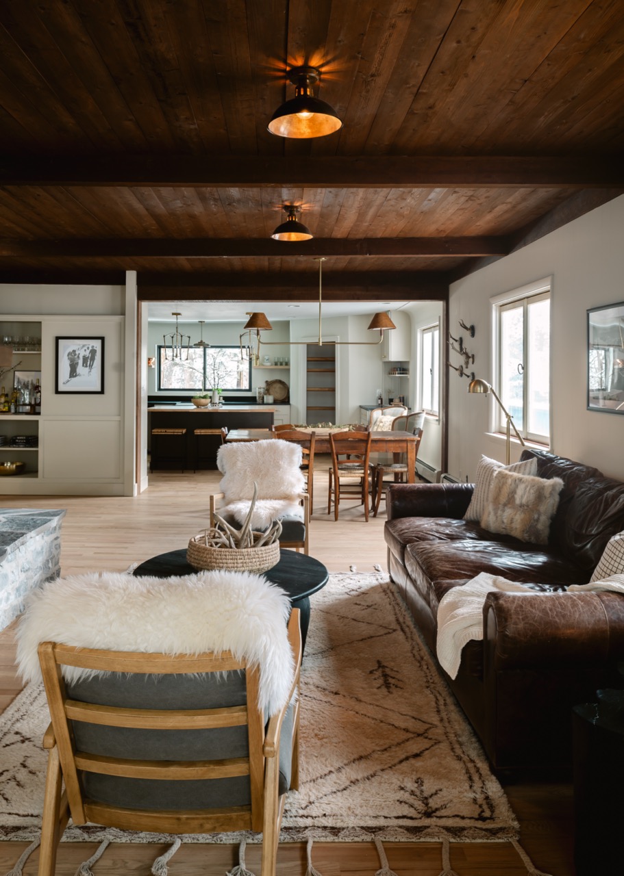 Cozy living room with wooden ceiling, leather sofa with pillows and throw, wooden chairs with white fur throws, a round coffee table, and a dining area with wooden chairs in the background.