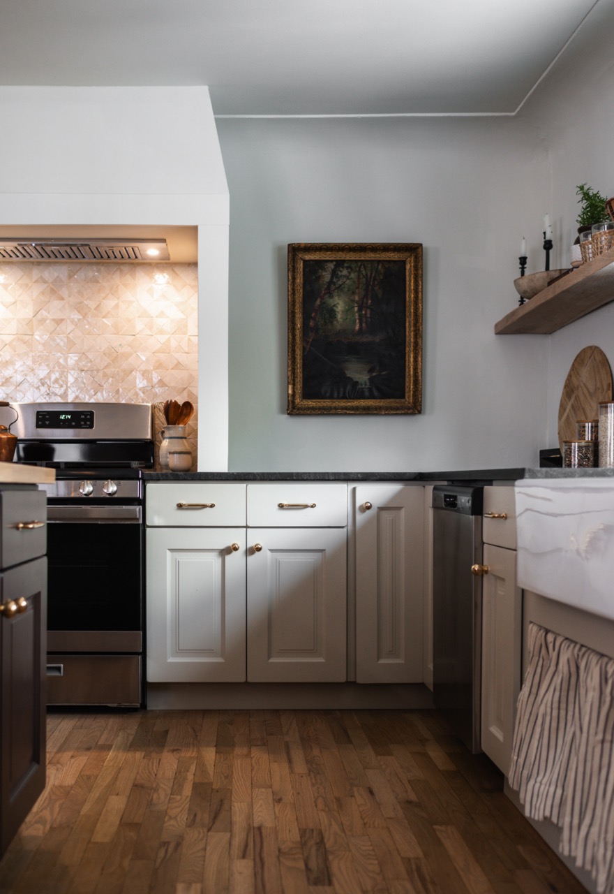 Modern kitchen with white cabinetry, black countertop, stainless steel stove, dishwasher, wooden floor, and framed painting on the wall.