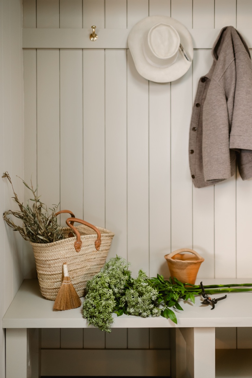 Cozy mudroom bench with wicker basket holding dried plants, small broom, fresh greenery, terracotta pot, pruning shears, beige coat and white hat hanging on wall hooks.