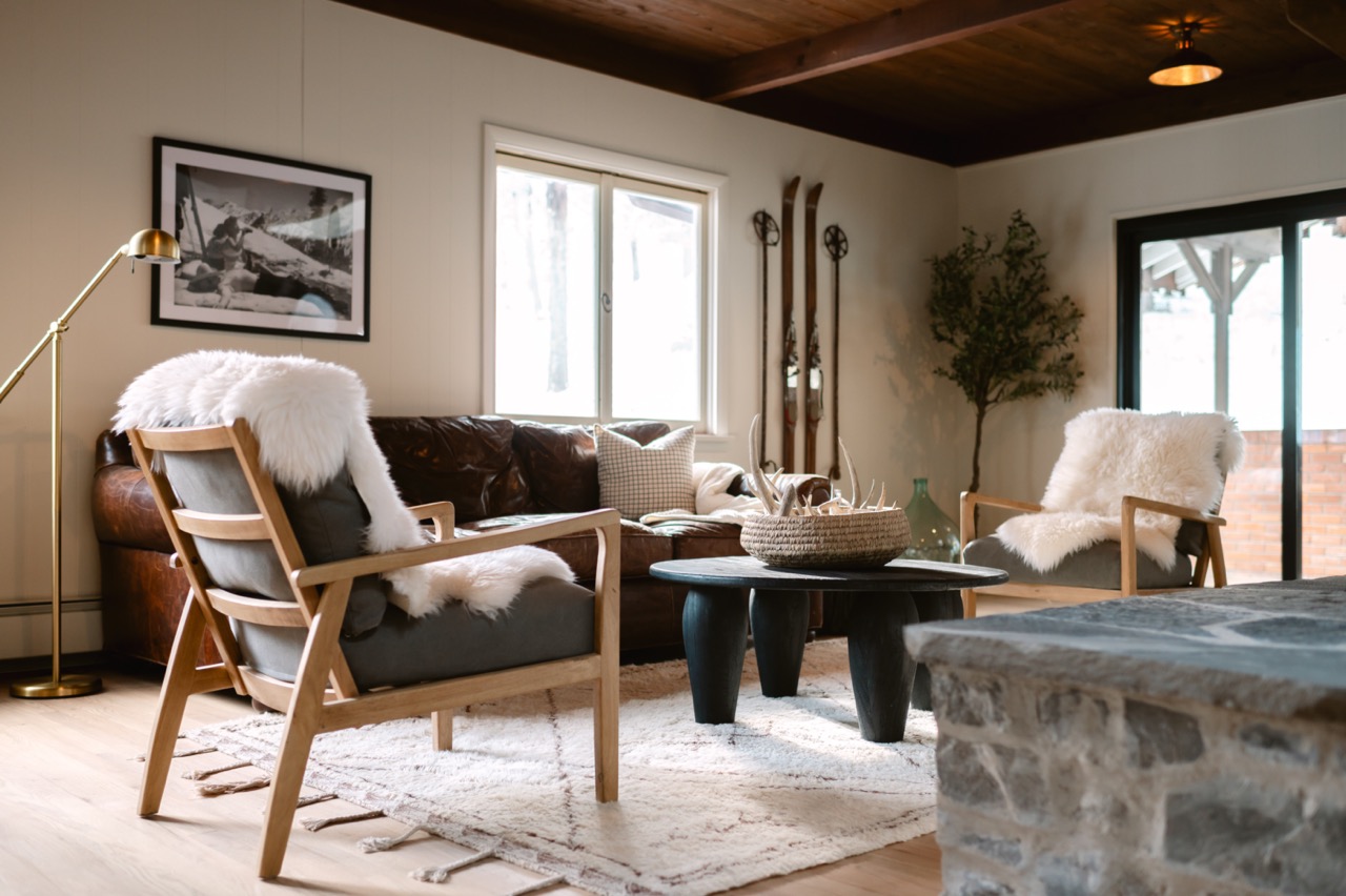 Cozy living room with wooden armchairs draped in white fur throws, a dark leather sofa, round black coffee table, and vintage skis on the wall.