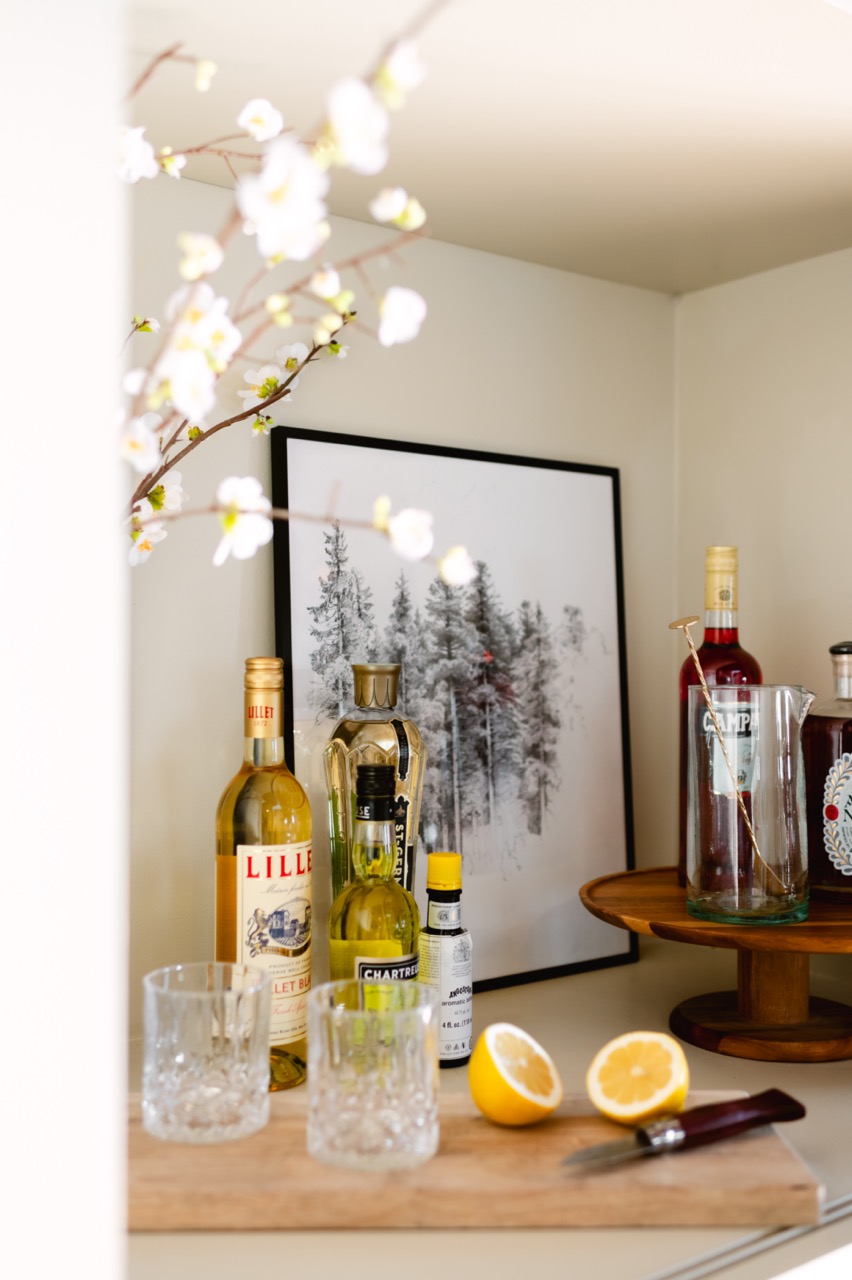 Countertop bar setup with bottles of liquor, two empty glasses, a halved lemon, and a framed black-and-white pine tree artwork in the background.