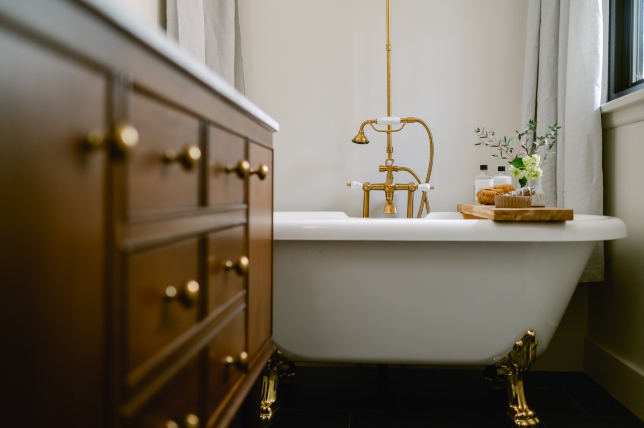 White clawfoot bathtub with brass fixtures and a wooden tray holding spa items beside a wooden cabinet with brass knobs.