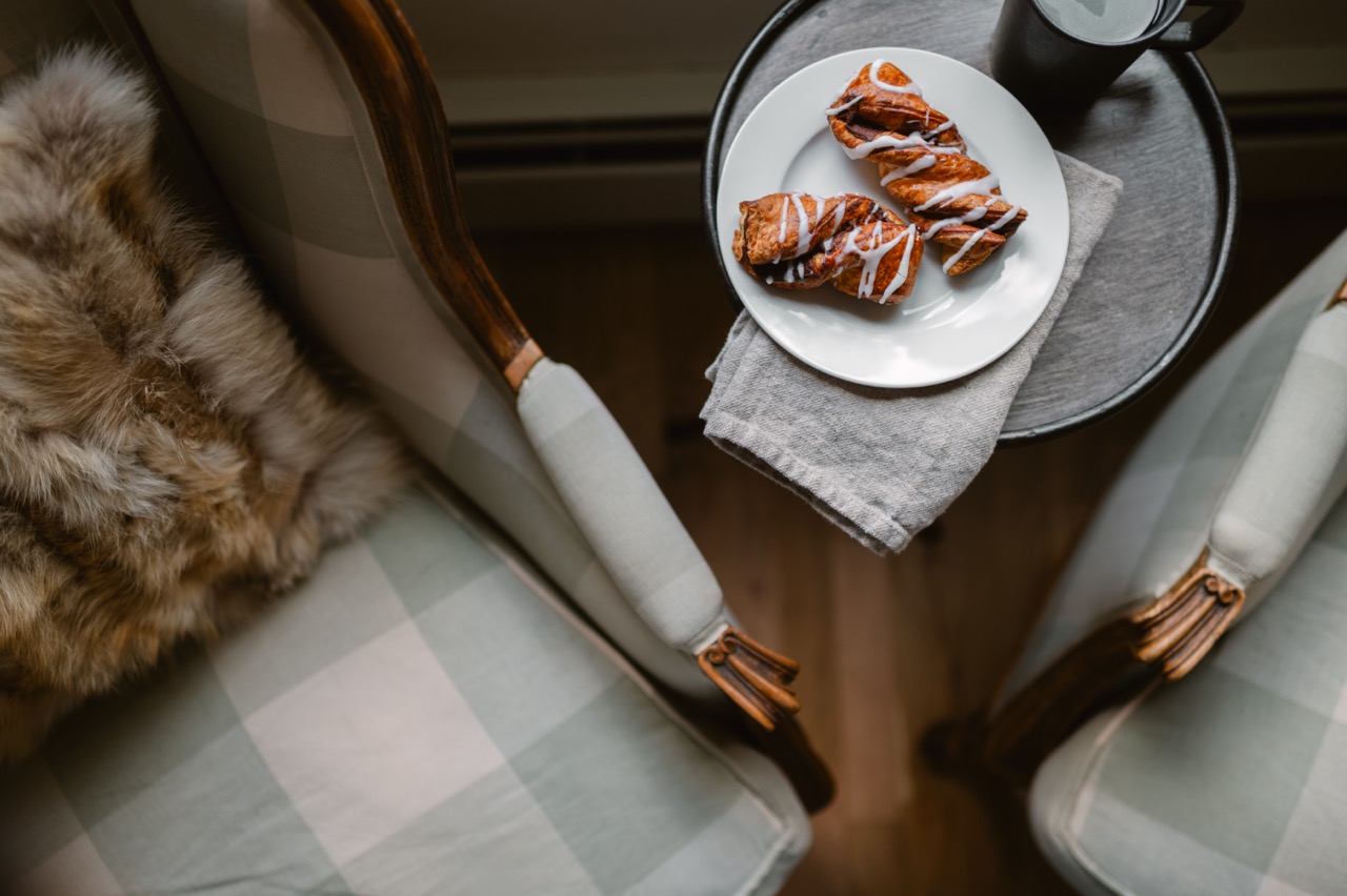 Two cinnamon twists with icing on a white plate next to a black cup on a round wooden table between two cushioned chairs.