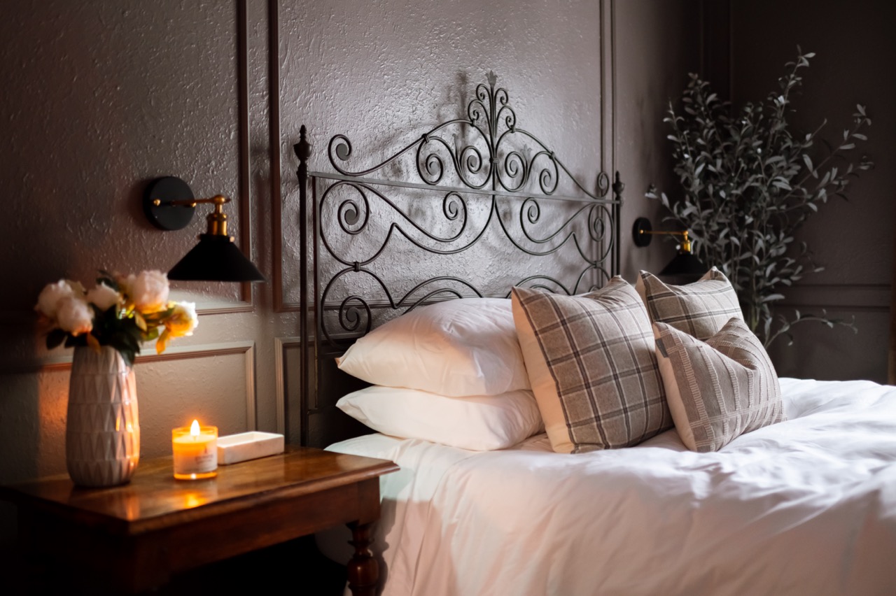 Cozy bedroom with a white bed, plaid pillows, ornate black metal headboard, wooden nightstand with lit candle and flower vase, and wall sconces.
