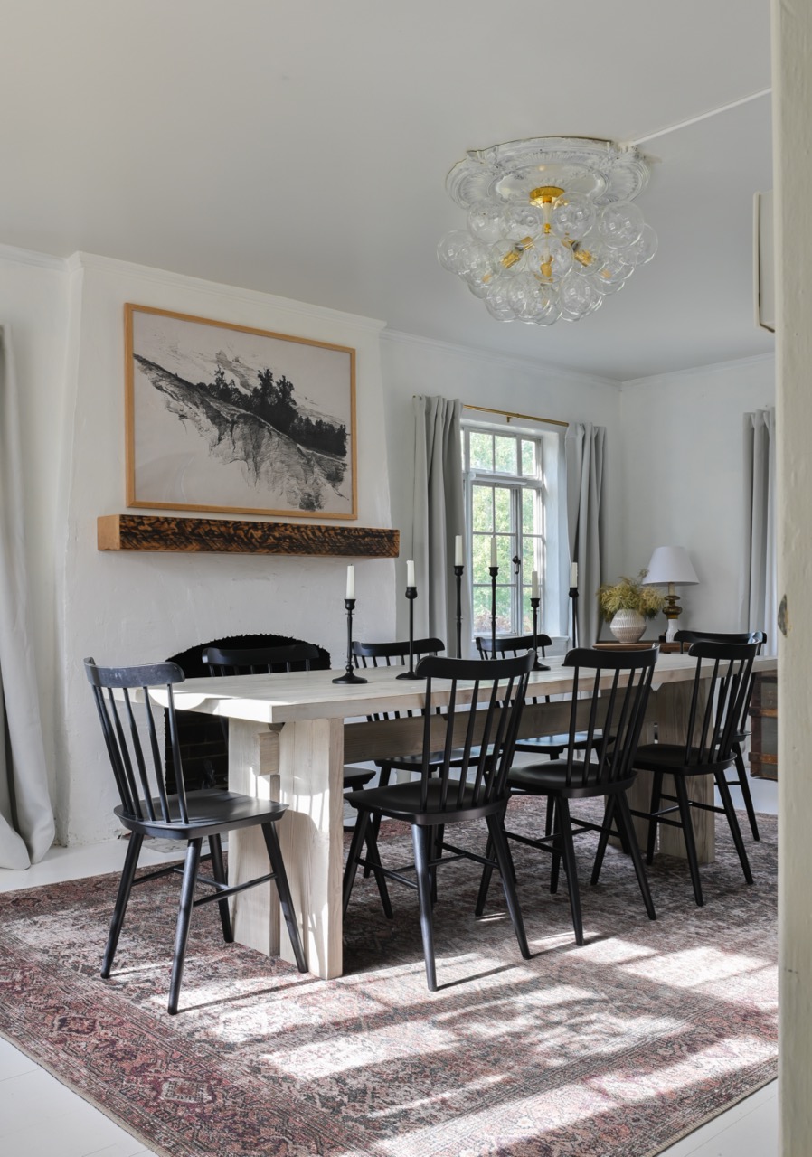 Dining room with a light wood table surrounded by black chairs, black candlesticks on the table, a patterned rug, and a window with gray curtains.