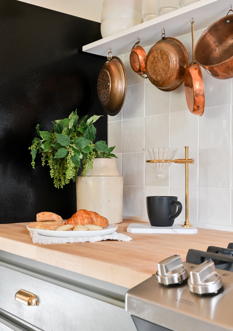 Kitchen countertop with a plate of pastries, a beige vase with green foliage, copper pots hanging above, and a black coffee cup under a brass coffee dripper.