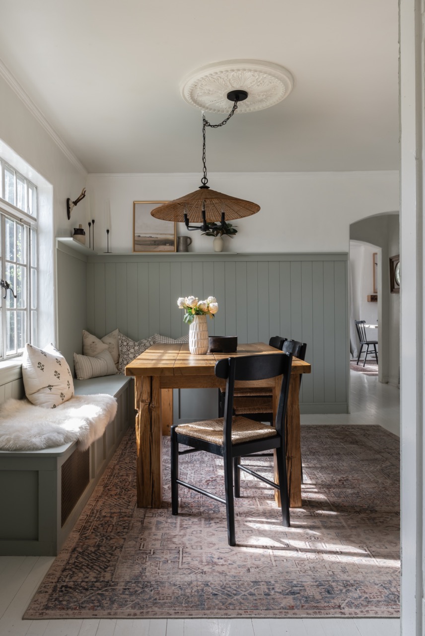 Dining nook with wooden table, black chairs, and built-in cushioned bench seating by a window, under a woven pendant light.