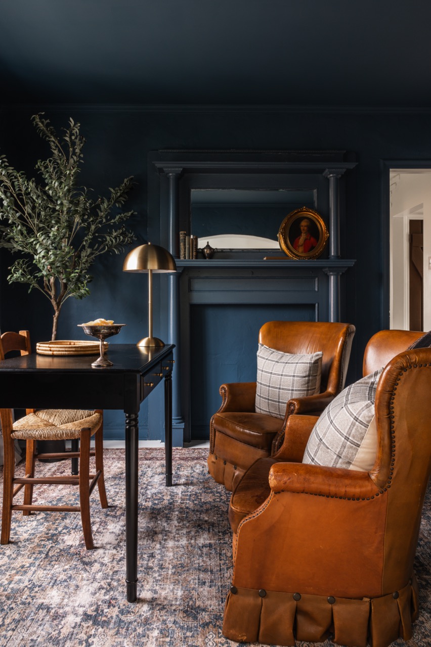 Cozy living room with dark blue walls, two brown leather armchairs with plaid pillows, a black table with a gold lamp, a wooden chair, and a decorative plant.