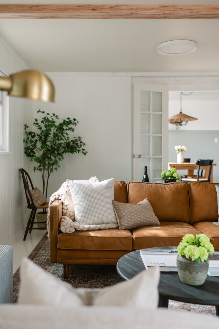 Cozy living room with a brown leather sofa adorned with cushions and a chunky knit blanket, a round black coffee table with a green plant, and a wooden chair with a pillow near a potted tree.