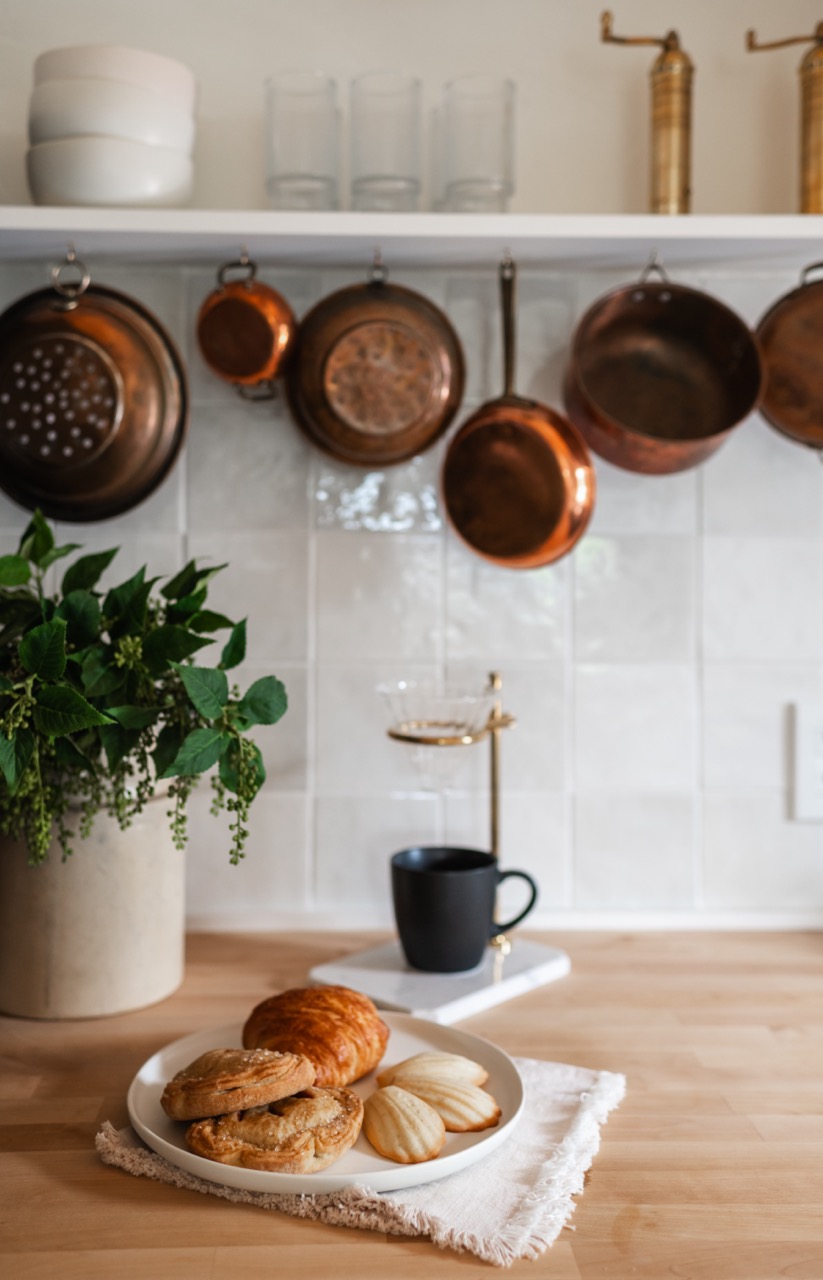 Plate with assorted pastries including a croissant and madeleines on a wooden countertop, with a black cup, green plant in a pot, and copper pans hanging on a white tiled kitchen wall.