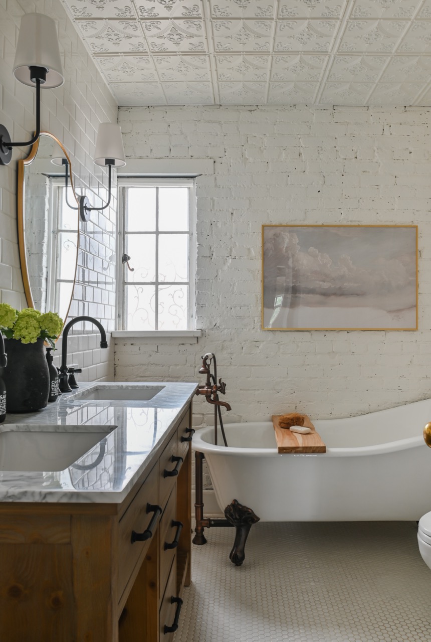 Bright bathroom with a white clawfoot bathtub, wooden vanity with marble countertop, round mirror, and white brick walls.