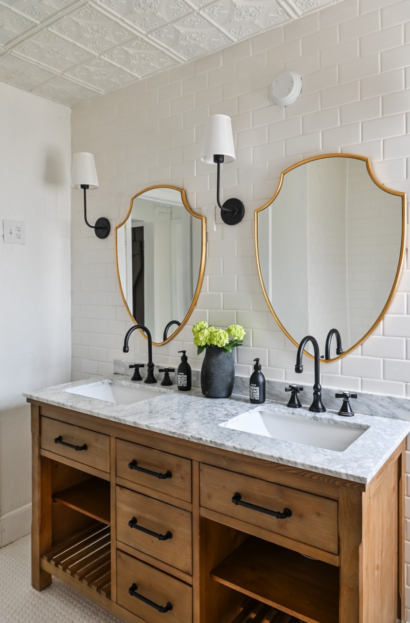 Double bathroom vanity with marble countertop, two gold-framed mirrors, black faucets, wall sconces, and a vase with green flowers.