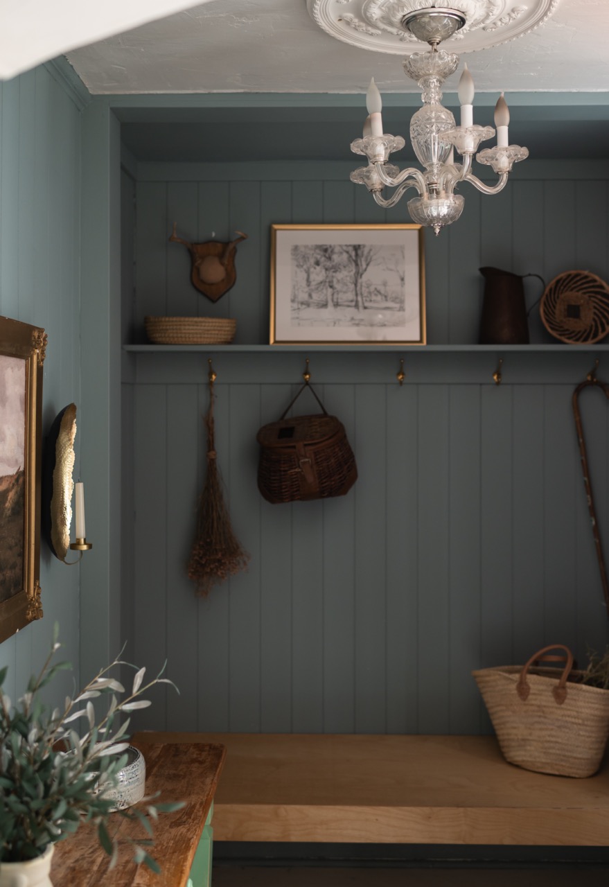 Cozy mudroom with blue-gray paneled walls, wooden bench, woven baskets, dried flowers, framed sketch, and a chandelier.