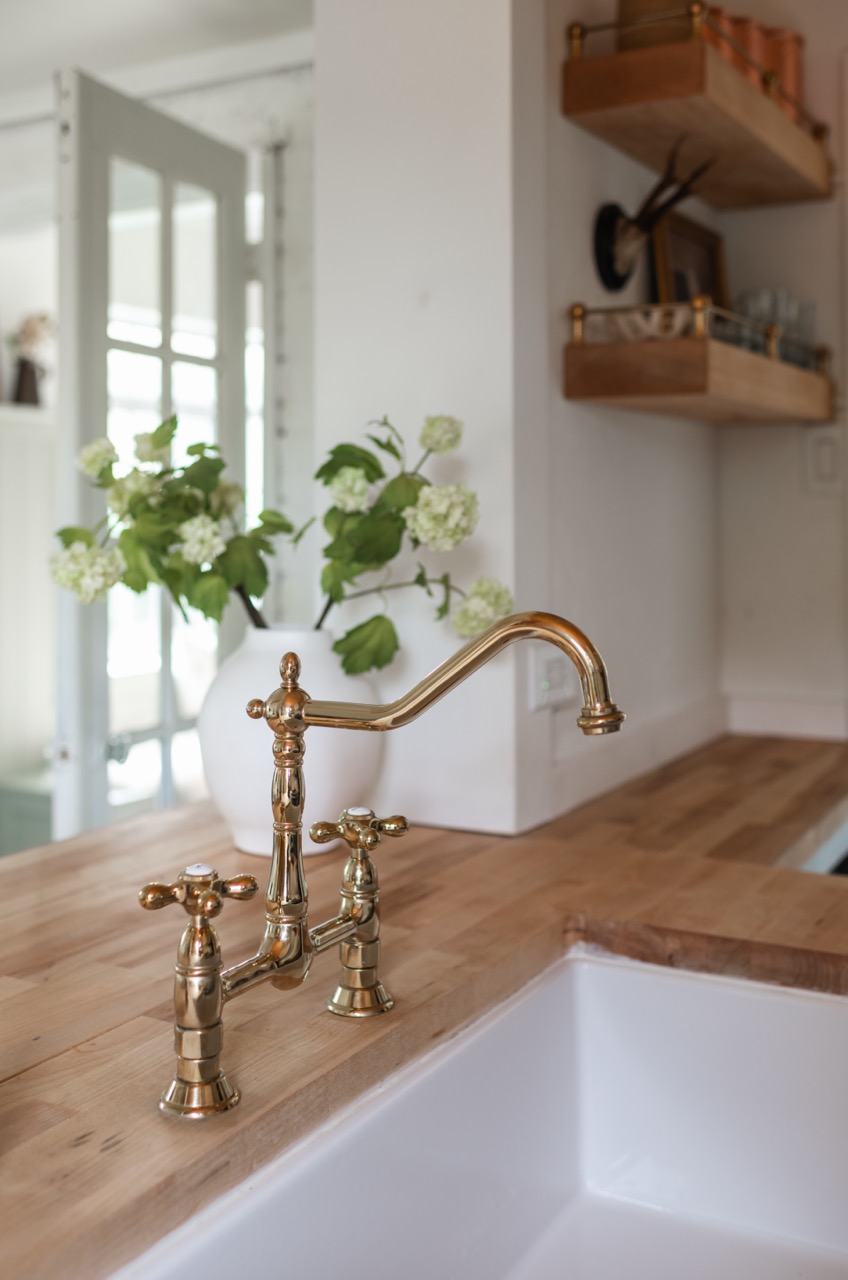 Vintage brass kitchen faucet mounted on wooden countertop next to white sink with a white vase of green and white flowers in the background.