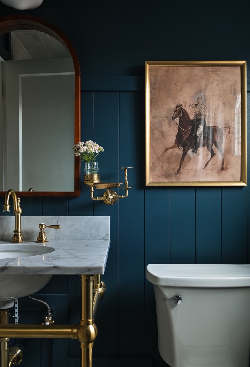 Bathroom with dark blue paneled wall, white marble sink with brass fixtures, a mounted brass vase holder with flowers, and framed painting of a horse and rider.