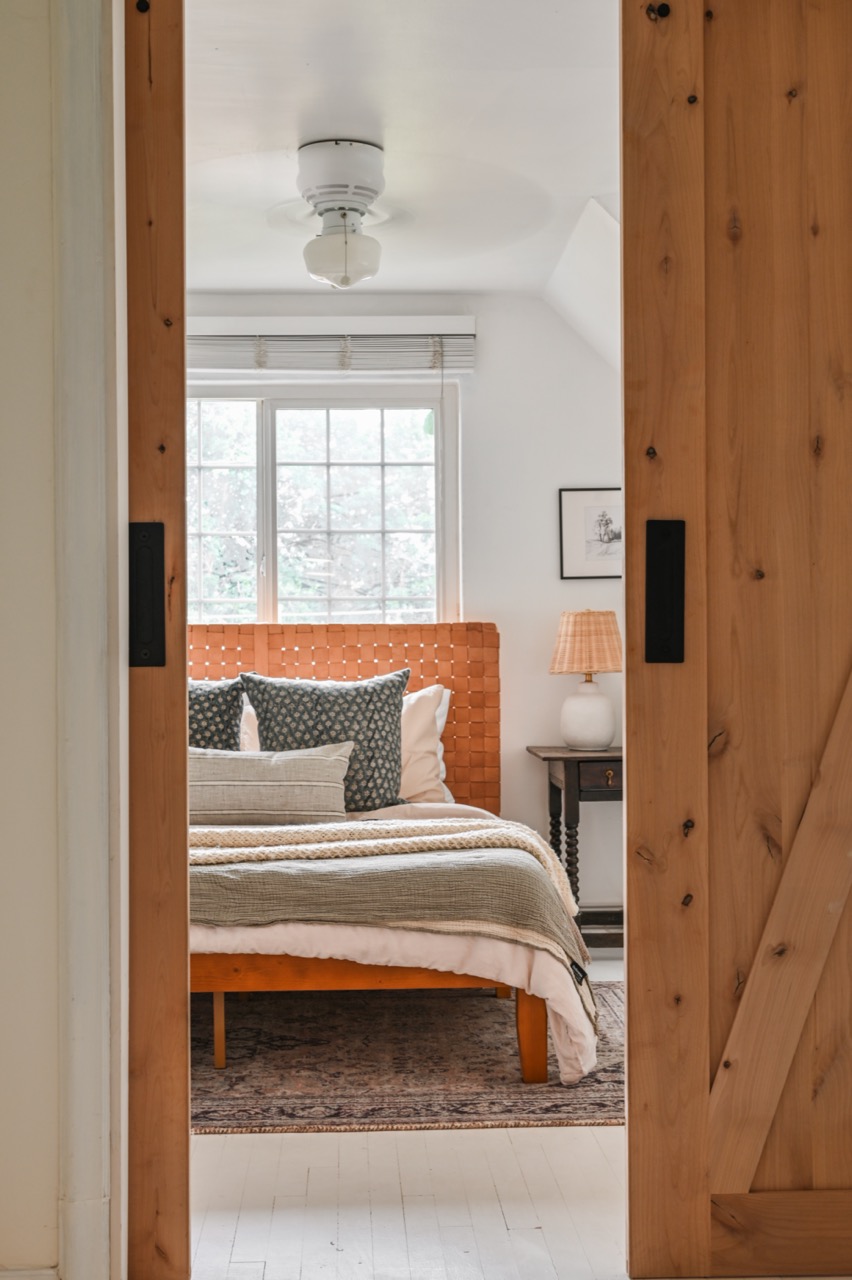 Cozy bedroom viewed through wooden sliding barn doors featuring a woven leather headboard, layered bedding, two patterned throw pillows, a bedside table with a lamp, and a large window with blinds.