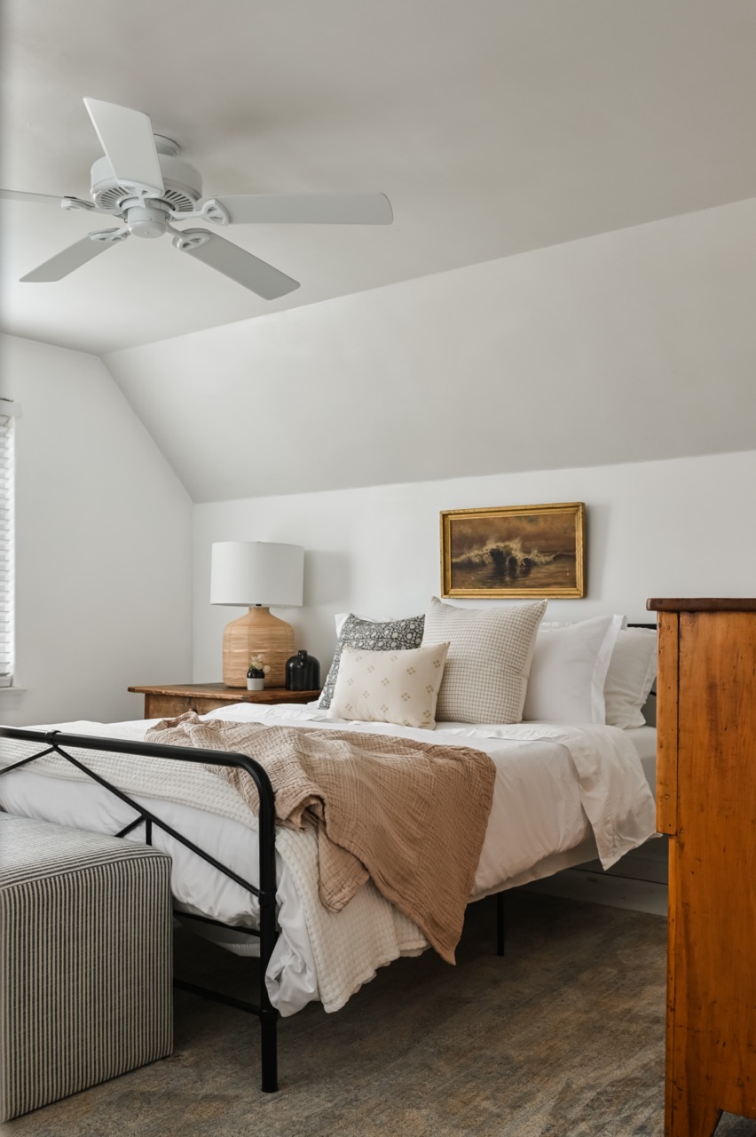 Cozy bedroom with a metal-framed bed, layered white and beige bedding, patterned pillows, wooden nightstand with lamp, striped ottoman, and a framed painting on white wall.