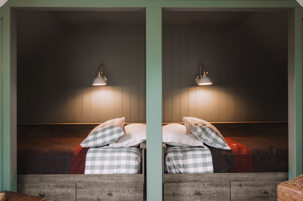 Two single beds with plaid pillows and blankets separated by a green divider, each under a wall-mounted reading lamp.