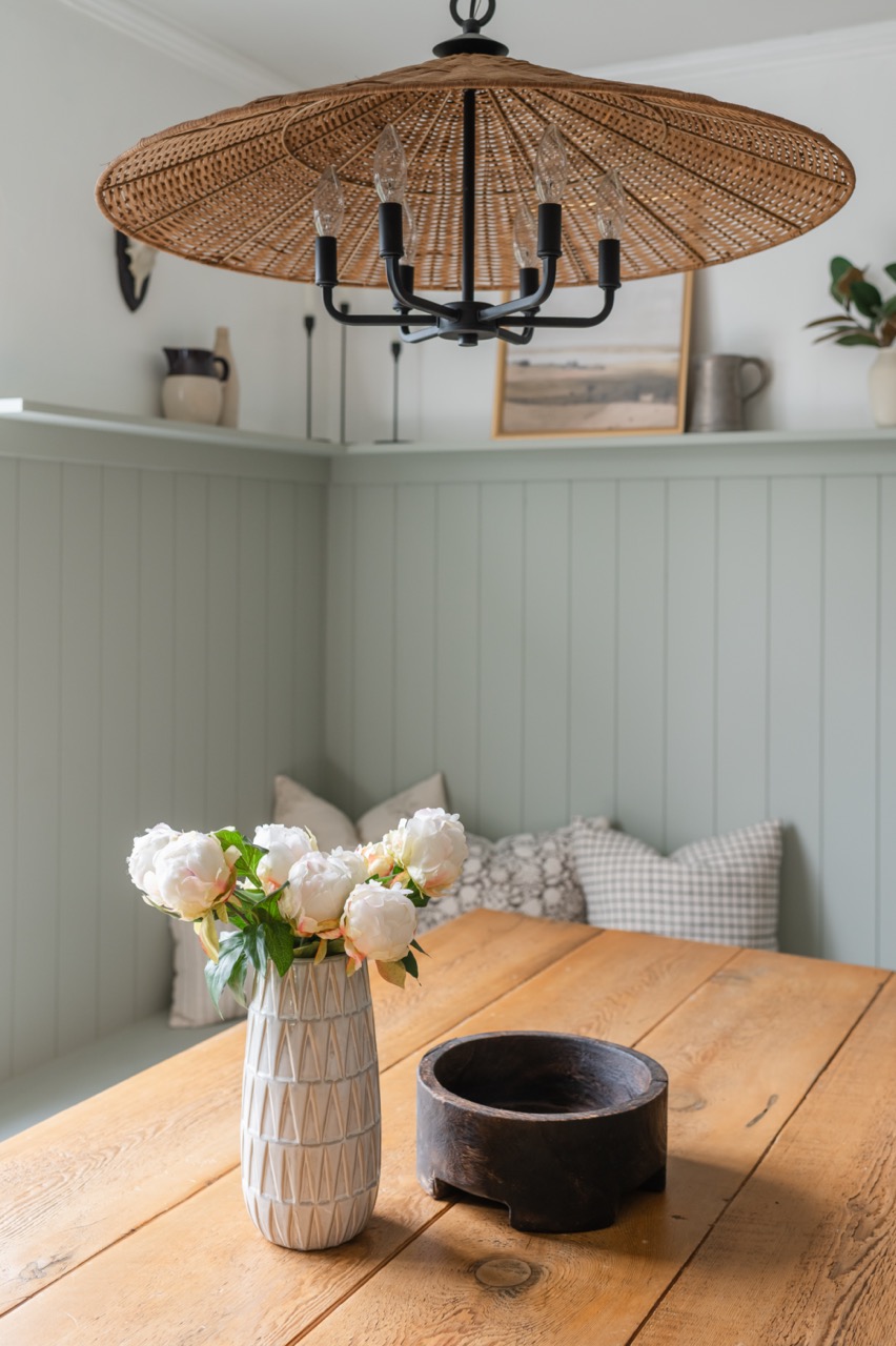 Wooden dining table with a white textured vase holding white flowers and a dark round wooden bowl under a large woven rattan chandelier in a cozy room with cushioned seating.