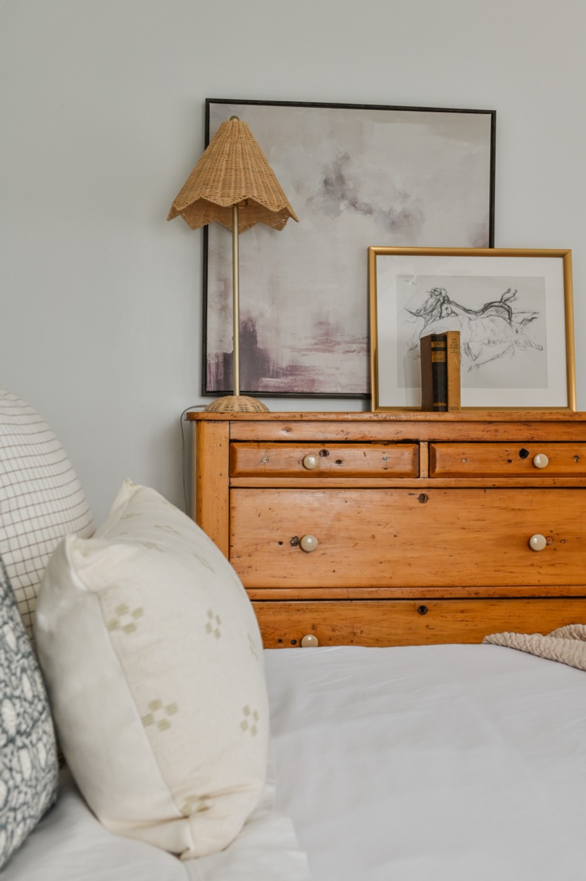 Wooden dresser with a woven lamp, framed abstract art, and a framed horse sketch, next to a bed with neutral patterned pillows.