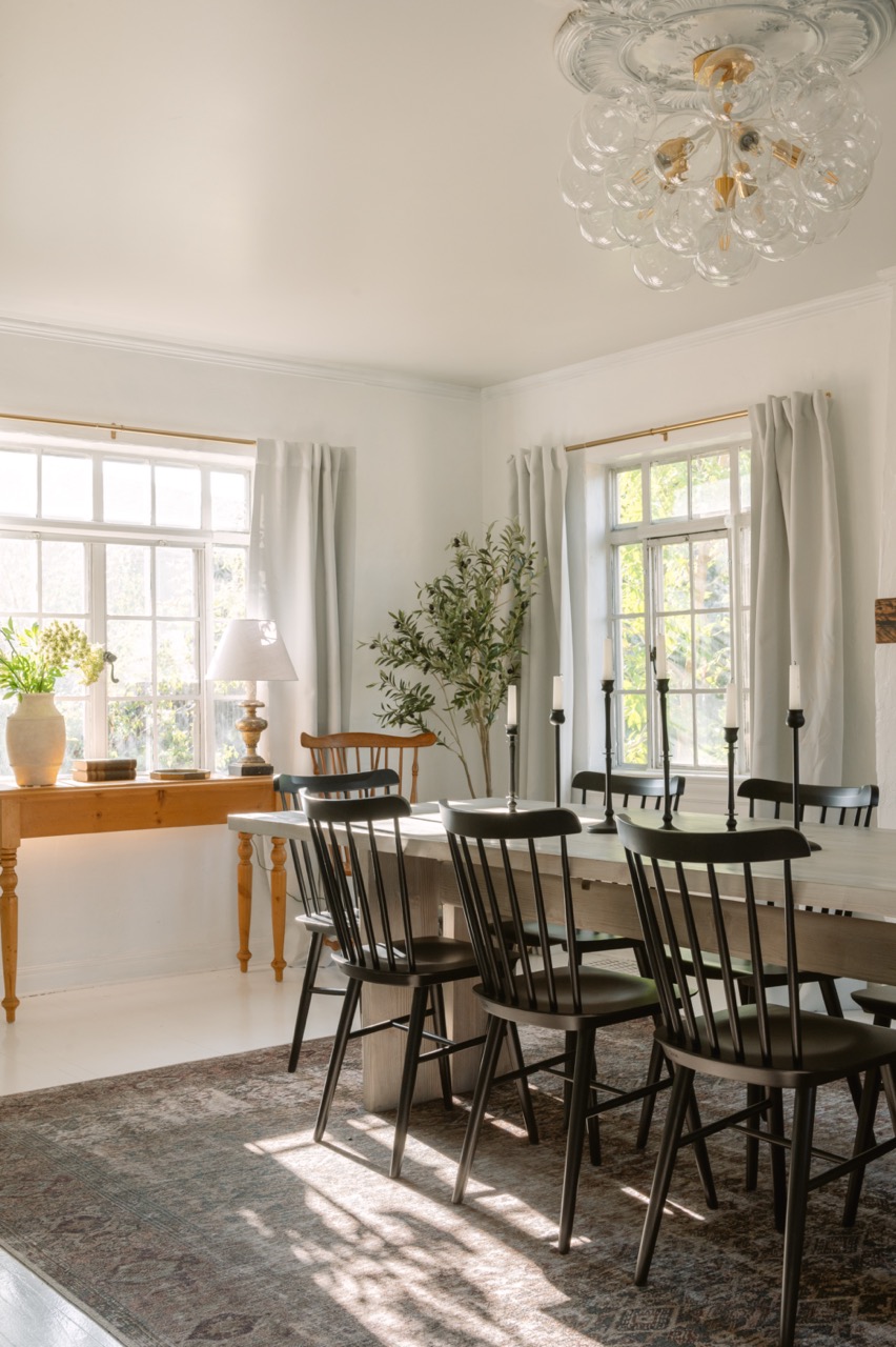 Bright dining room with black chairs around a wooden table, large windows with curtains, a wooden side table with a lamp and vase, and a decorative chandelier.