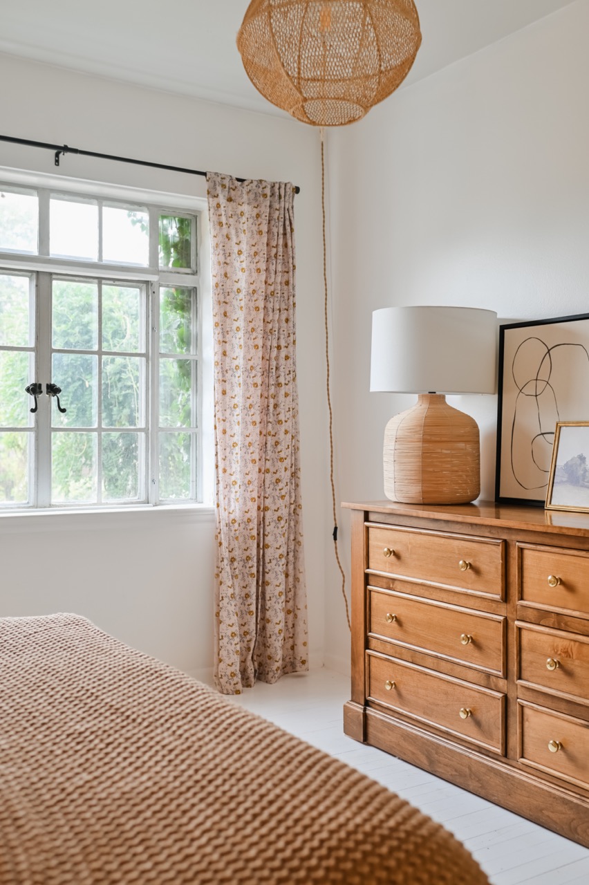 Cozy bedroom corner with a wooden dresser topped with a woven lamp and abstract art, beside a window with floral curtains and a textured brown blanket.