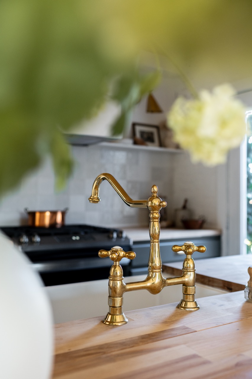 Brass kitchen faucet mounted on a wooden countertop with a stove and white tiled backsplash in the background.