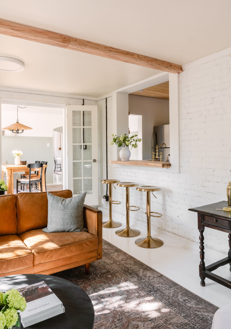 Bright living room with tan leather sofa, gray pillow, brass bar stools at a white brick wall, and a window opening to the kitchen.