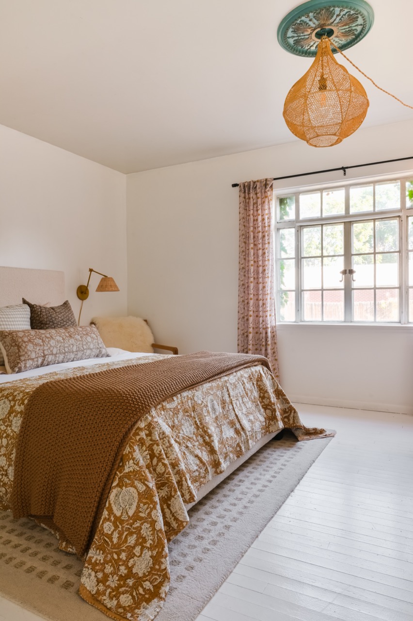 Cozy bedroom with a patterned brown and white bedspread, knitted blanket, patterned pillows, a window with floral curtains, and a woven pendant light.